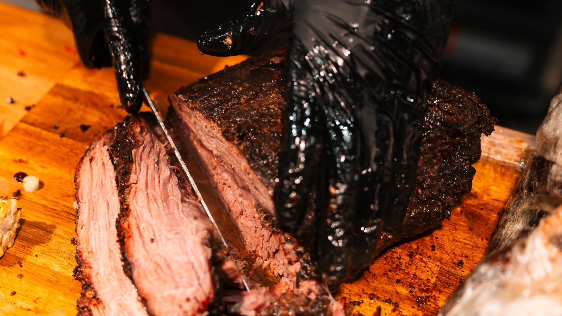 Pitmaster in black gloves slicing a smoked beef brisket with a visible smoke ring on a wooden board at The Backyard Smokehouse