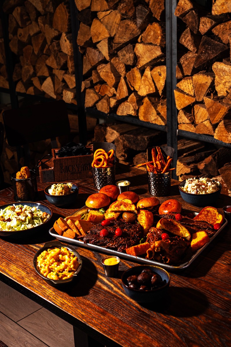 Overhead view of a full Backyard Smokehouse BBQ feast with smoked meats and sides on a wooden table against a stacked firewood wall