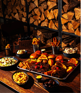 Diverse BBQ feast with sliders, brisket, and sides stacked near a woodpile at Edmonton location.