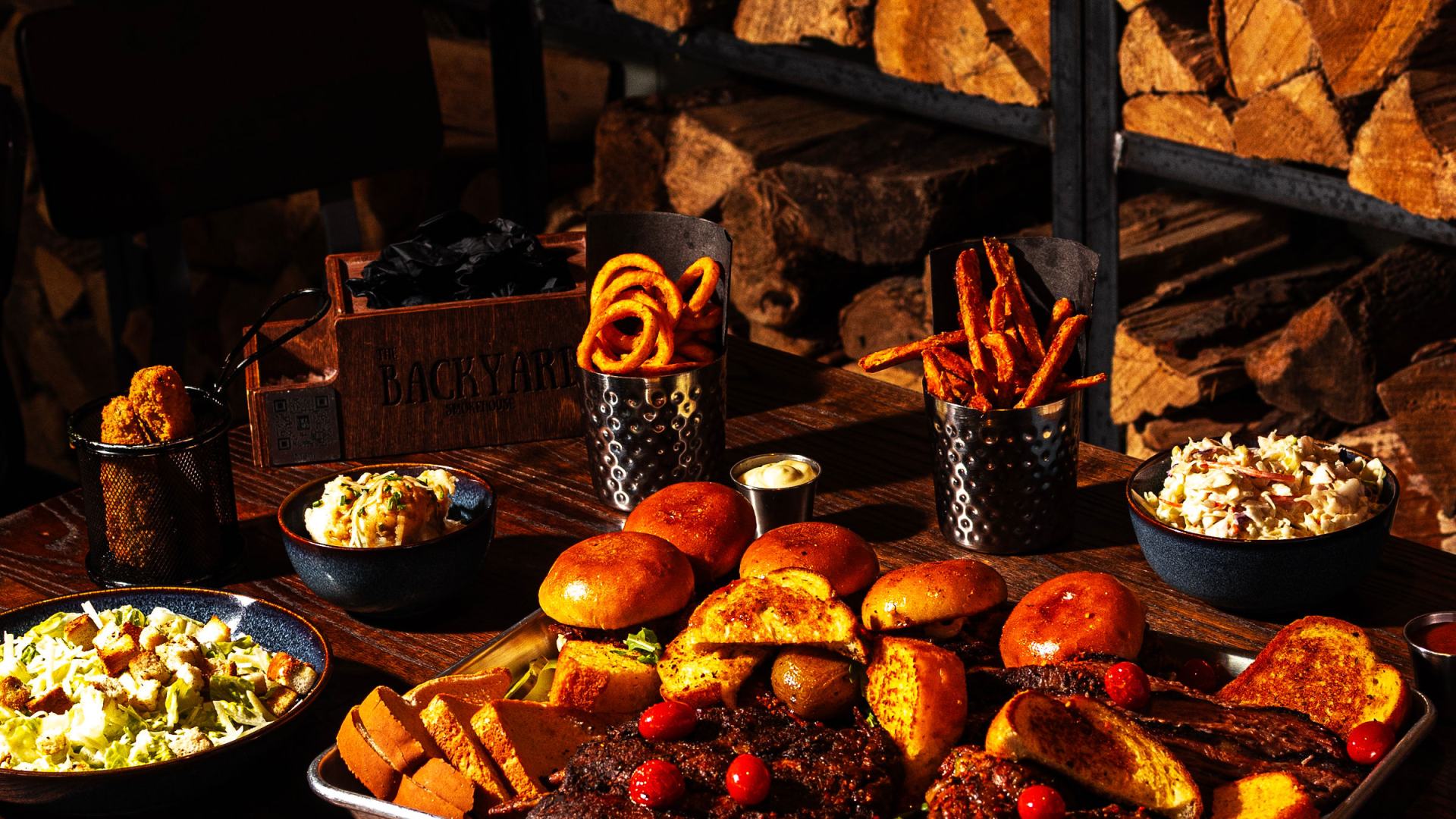The Backyard Smokehouse feast spread with smoked meats, mac and cheese, coleslaw and curly fries on a wooden table by a stacked firewood wall