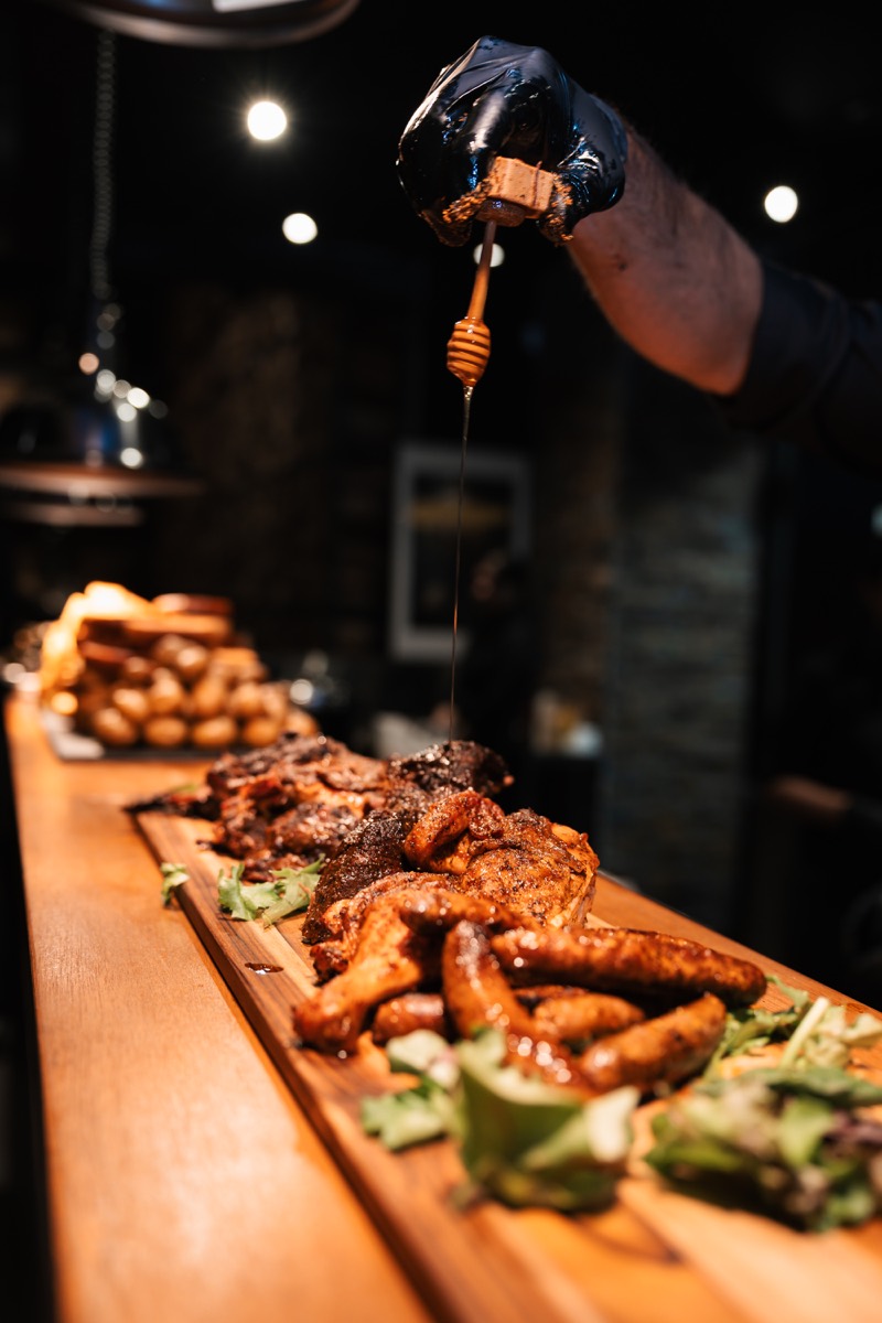 Honey drizzled over a long wooden board of grilled chicken, sausages and smoked meats at The Backyard Smokehouse